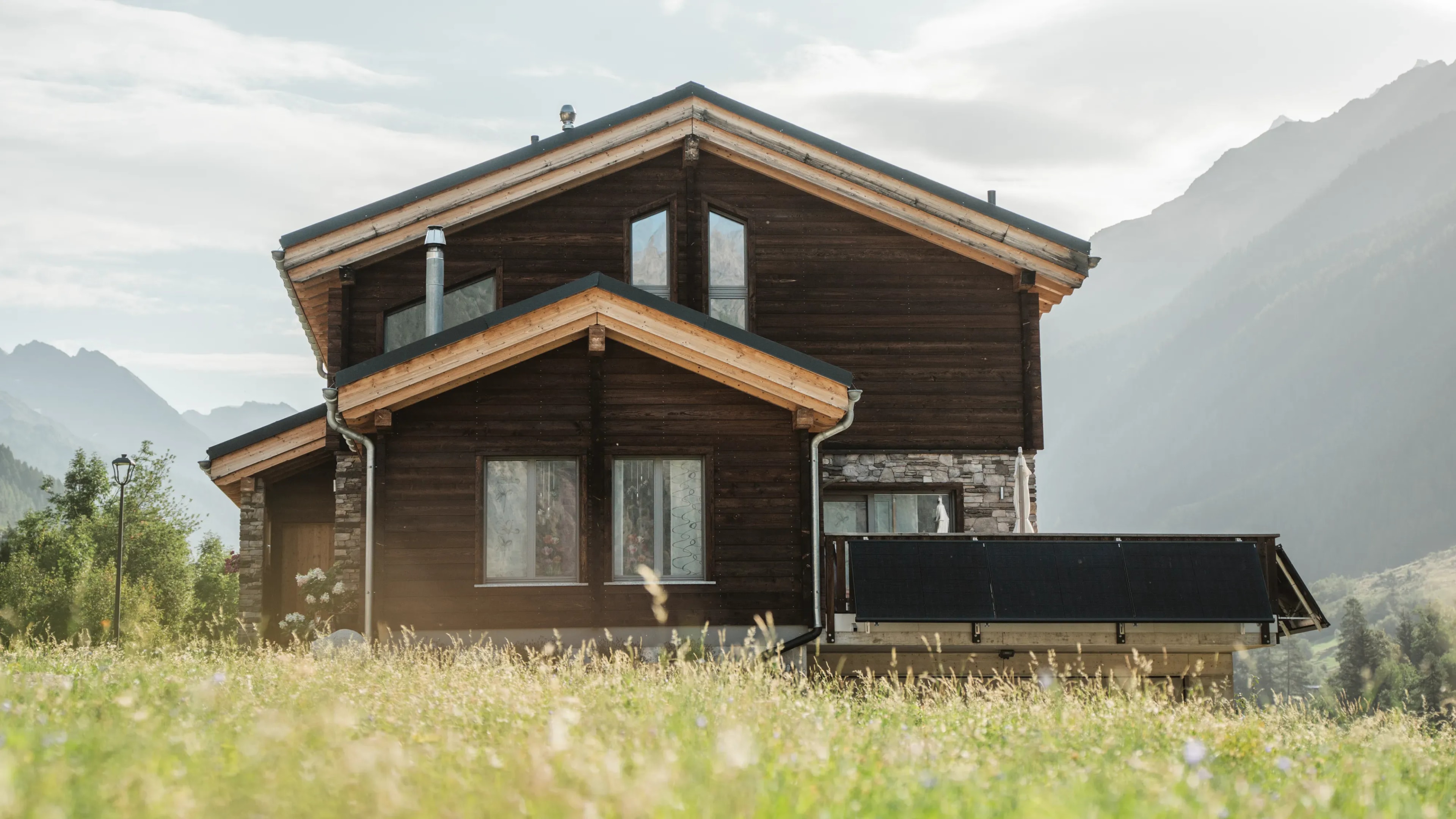 Chalet mit dunkler Holzfassade, Satteldächern und Solarpanels am Balkon vor Berglandschaft, im Vordergrund eine Blumenwiese.