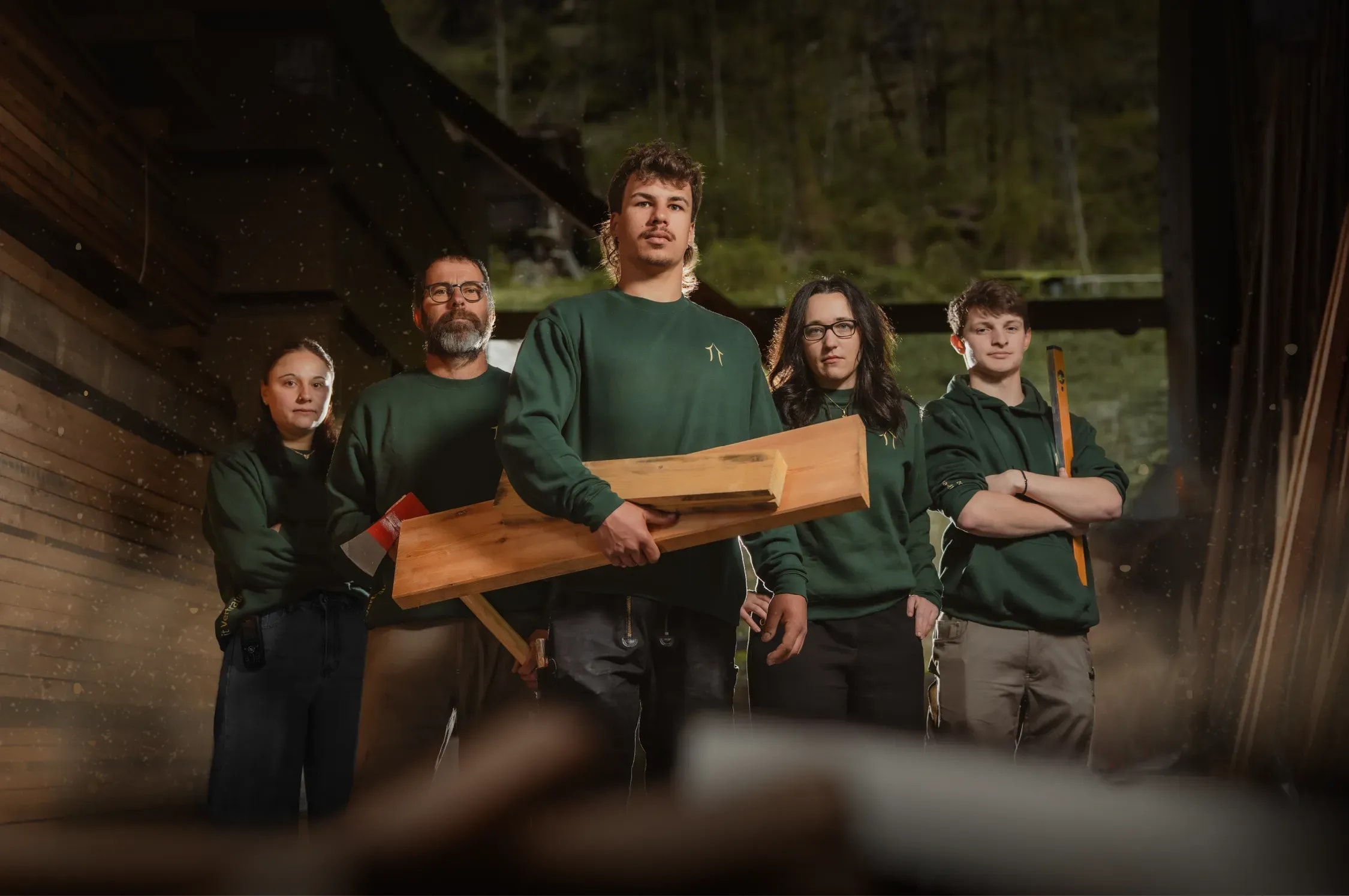 Team der Holzbau AG in grünen Firmenpullovern mit Holzbalken, Axt und Wasserwaage in einer Werkhalle, Kampagnenfoto von Schnyder Werbung.
