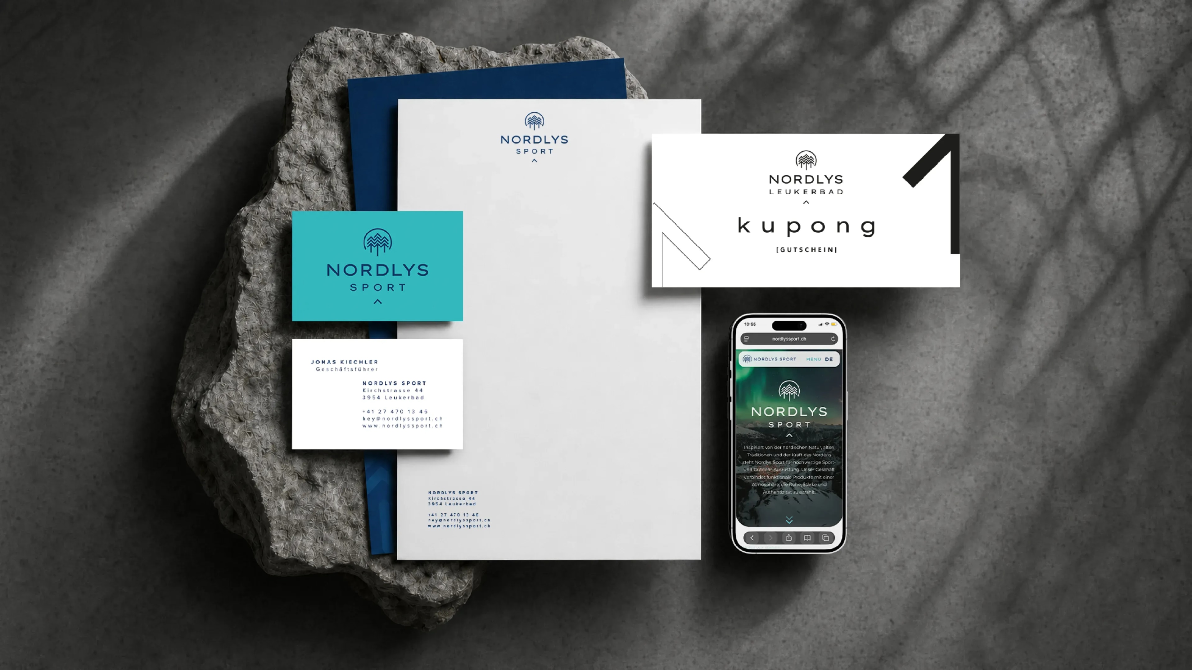 Geschäftsausstattung von Nordlys Sport auf einem Stein: Briefpapier, Visitenkarten in Türkis und Weiss, Gutschein ‚kupong' und Website auf dem Smartphone.