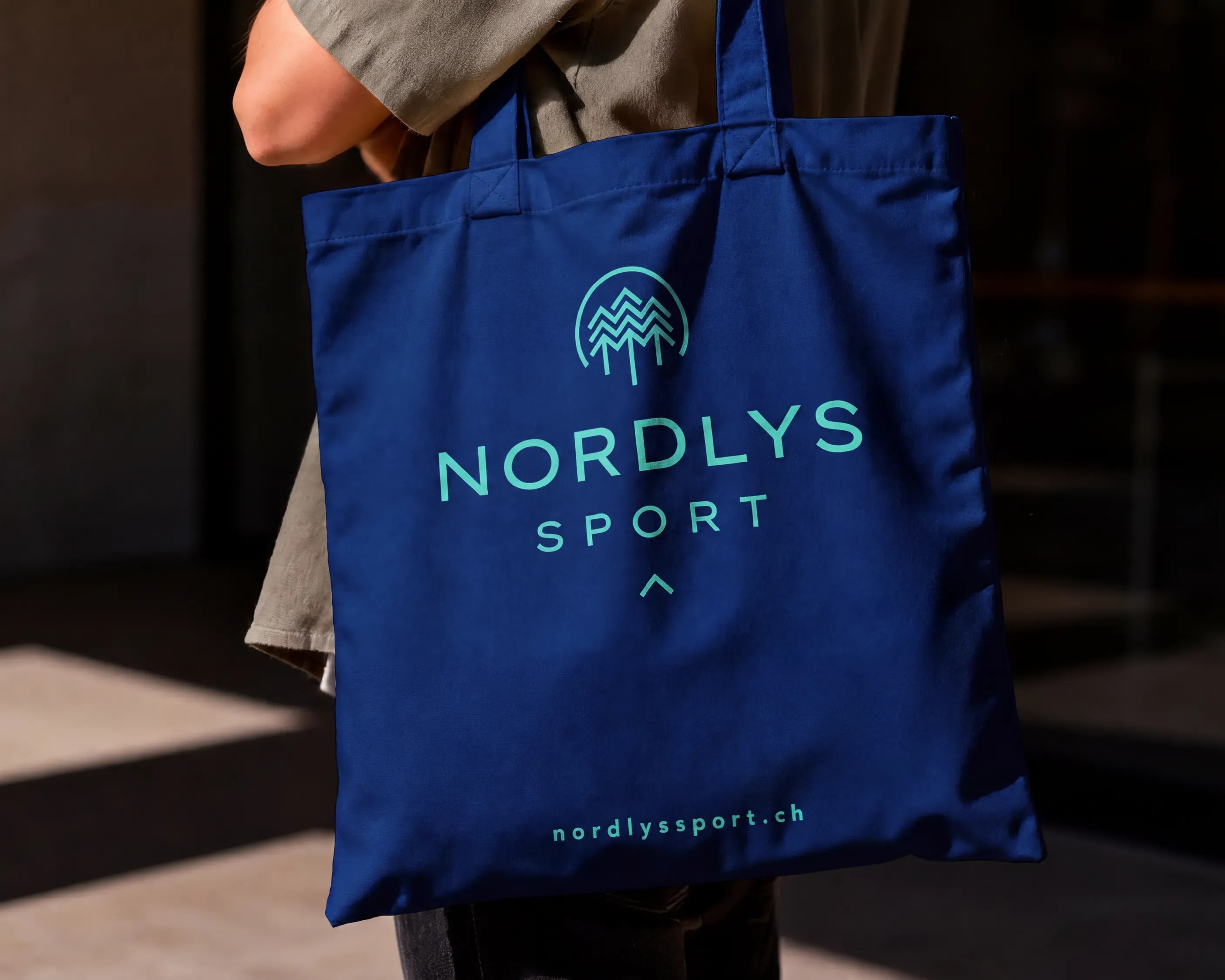 Person trägt eine blaue Stofftasche mit türkisem Nordlys-Sport-Logo und der Adresse nordlyssport.ch.