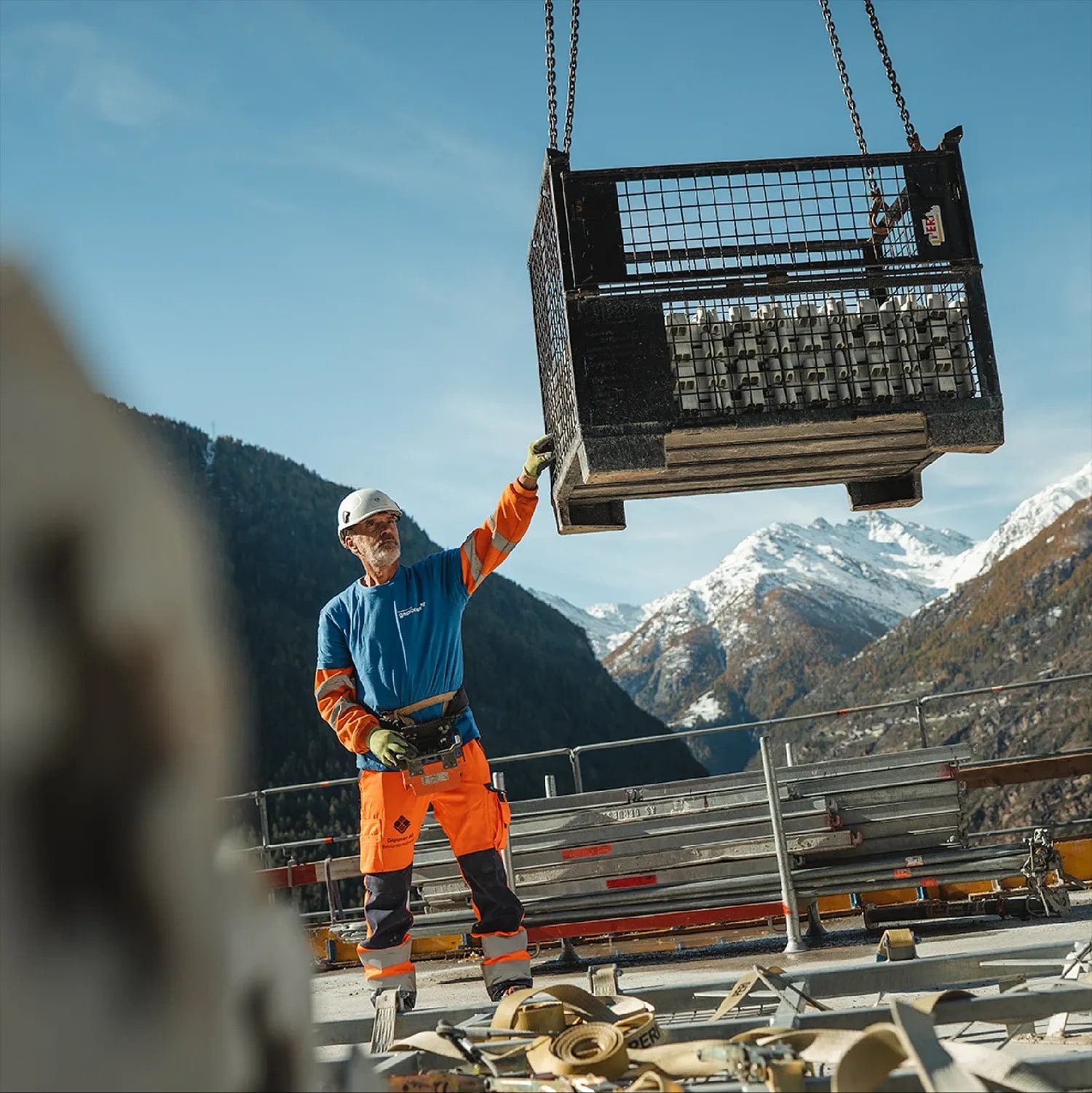 Bauarbeiter in Warnkleidung führt eine am Kran hängende Gitterbox, dahinter verschneite Berge.