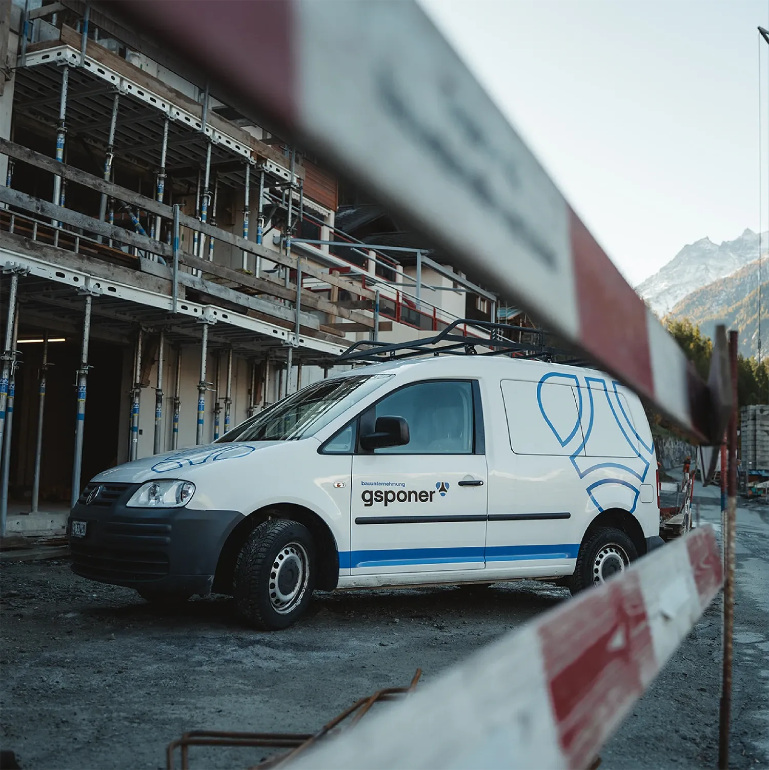 Beschrifteter Lieferwagen der Bauunternehmung Gsponer vor einem eingerüsteten Rohbau, im Vordergrund eine Absperrlatte.